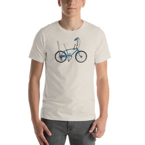 Peut inclure: Un t-shirt blanc avec un vélo bleu clair avec une selle banane et un guidon chromé.