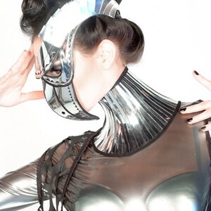 May include: A woman wearing a silver and black futuristic costume with a mirrored collar and a helmet-like mask. The costume has a mesh-like design on the shoulders and chest.