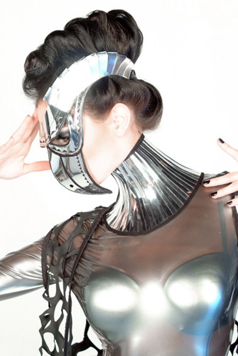 May include: A woman wearing a silver and black futuristic costume with a mirrored collar and a helmet-like mask. The costume has a mesh-like design on the shoulders and chest.