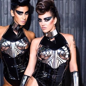May include: Two women wearing black and silver futuristic-style corsets with silver accents. The women have dark hair and dramatic eye makeup.