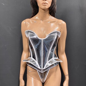 May include: A silver and white corset with a white trim and a pointed bottom. The corset is made of a shiny, reflective material.