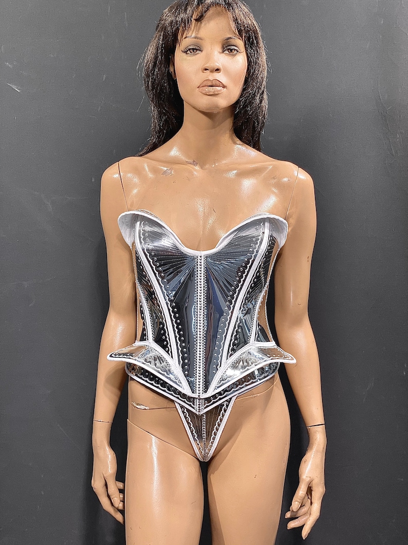 May include: A silver and white corset with a white trim and a pointed bottom. The corset is made of a shiny, reflective material.