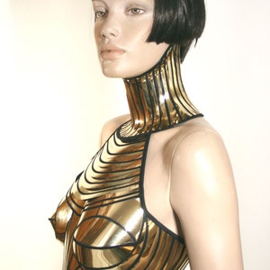 May include: A gold-coloured metallic chest piece with a high neck collar, featuring horizontal and vertical paneling with black trim. The garment covers the upper torso and neck, constructed from reflective, rigid segments that contour to the body.
