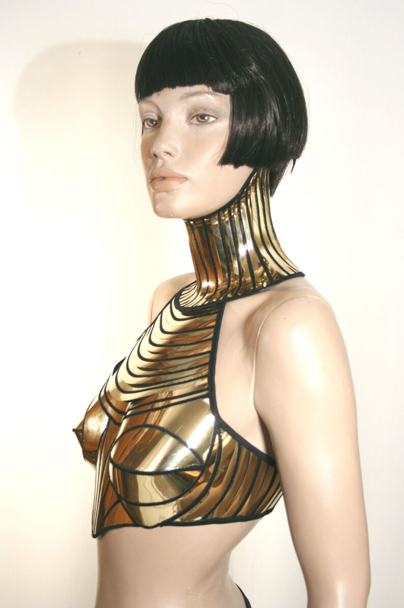 May include: A gold-coloured metallic chest piece with a high neck collar, featuring horizontal and vertical paneling with black trim. The garment covers the upper torso and neck, constructed from reflective, rigid segments that contour to the body.