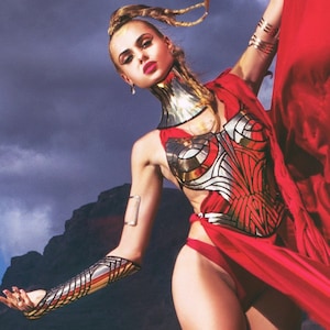 May include: A woman wearing a red dress and silver and gold armor. The armor is detailed and intricate, with a high collar and a bodice. The woman is posing with her arms outstretched, and her hair is pulled back in a ponytail.