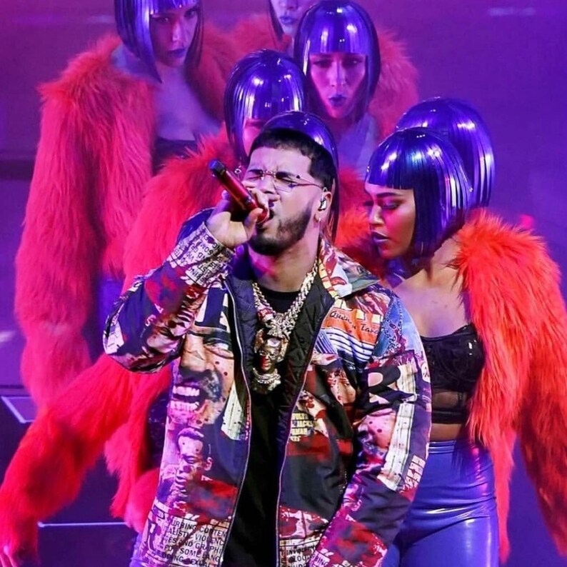 May include: A man wearing a colorful patterned jacket with a gold chain necklace sings into a microphone. He is surrounded by women wearing red fur jackets and purple wigs.