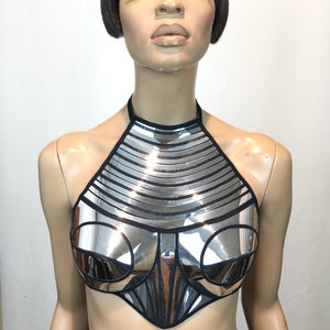 May include: A silver metallic halter top featuring a reflective, segmented panel design with black trim. The garment has a high neckline, structured circular chest panels, and a pointed hemline, creating a rigid, armour-like appearance.