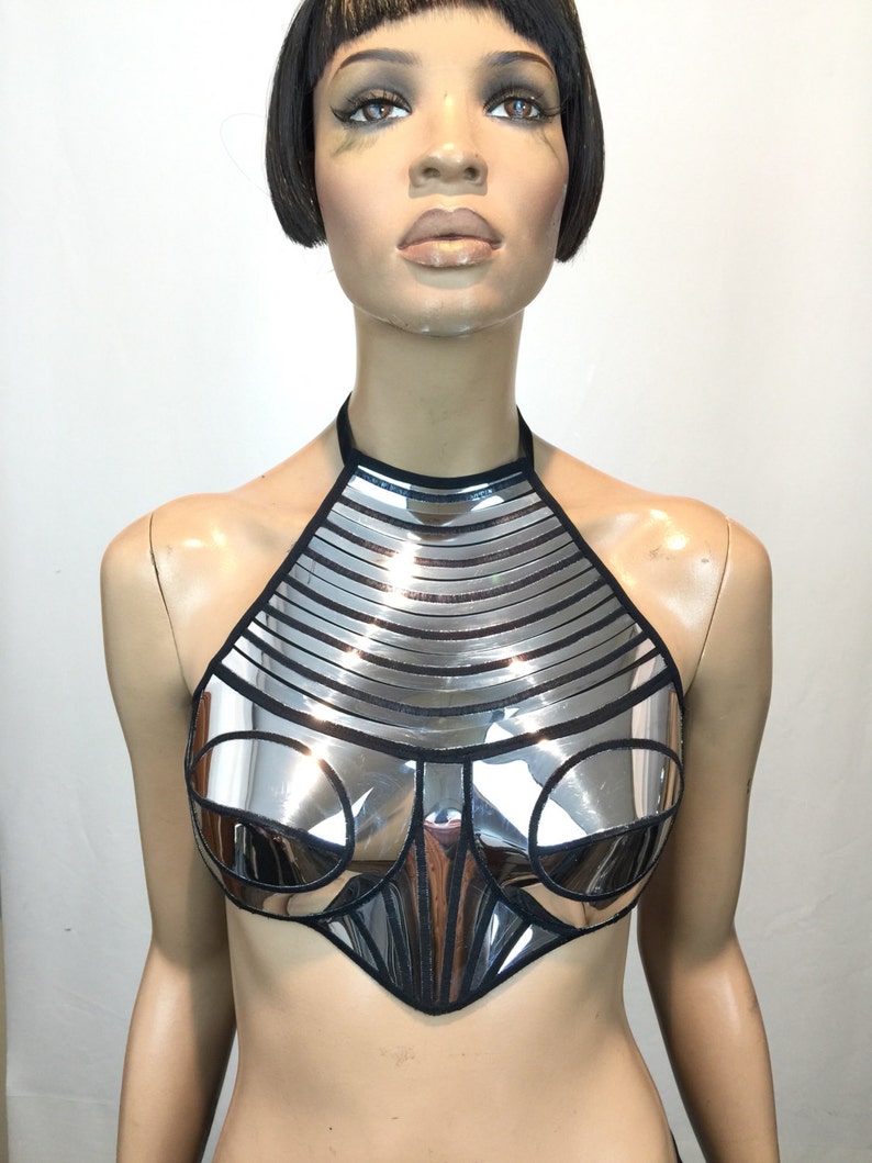 May include: A silver metallic halter top featuring a reflective, segmented panel design with black trim. The garment has a high neckline, structured circular chest panels, and a pointed hemline, creating a rigid, armour-like appearance.