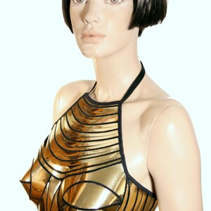 May include: A metallic gold halter top constructed from segmented, reflective panels with black trim. The garment features a structured, moulded bodice design with a high neckline and thin black straps that secure around the neck and back.