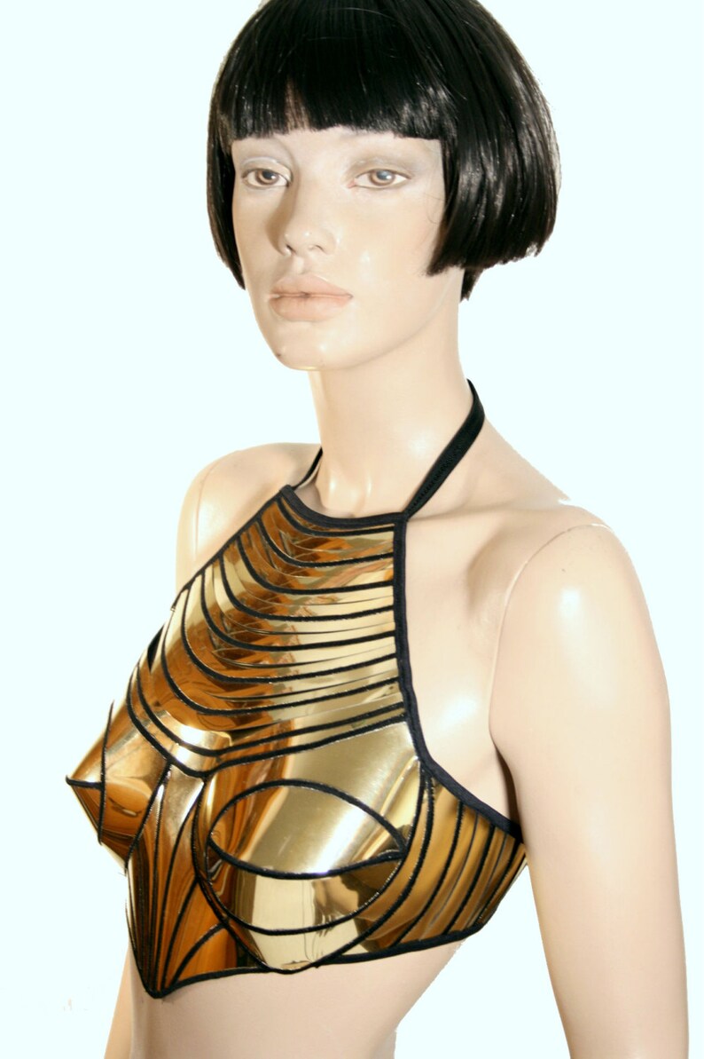 May include: A metallic gold halter top constructed from segmented, reflective panels with black trim. The garment features a structured, moulded bodice design with a high neckline and thin black straps that secure around the neck and back.