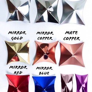 Pu&ograve; includere: Dodici piccole piastrelle metalliche decorative di forma quadrata, in vari colori e finiture. Le piastrelle sono disposte in tre file di quattro. La fila superiore presenta cromo a specchio, cromo scuro e argento opaco. La seconda fila presenta oro a specchio, rame a specchio e rame opaco. La terza fila presenta rosso a specchio, blu a specchio, rosa metallizzato e magenta metallizzato.