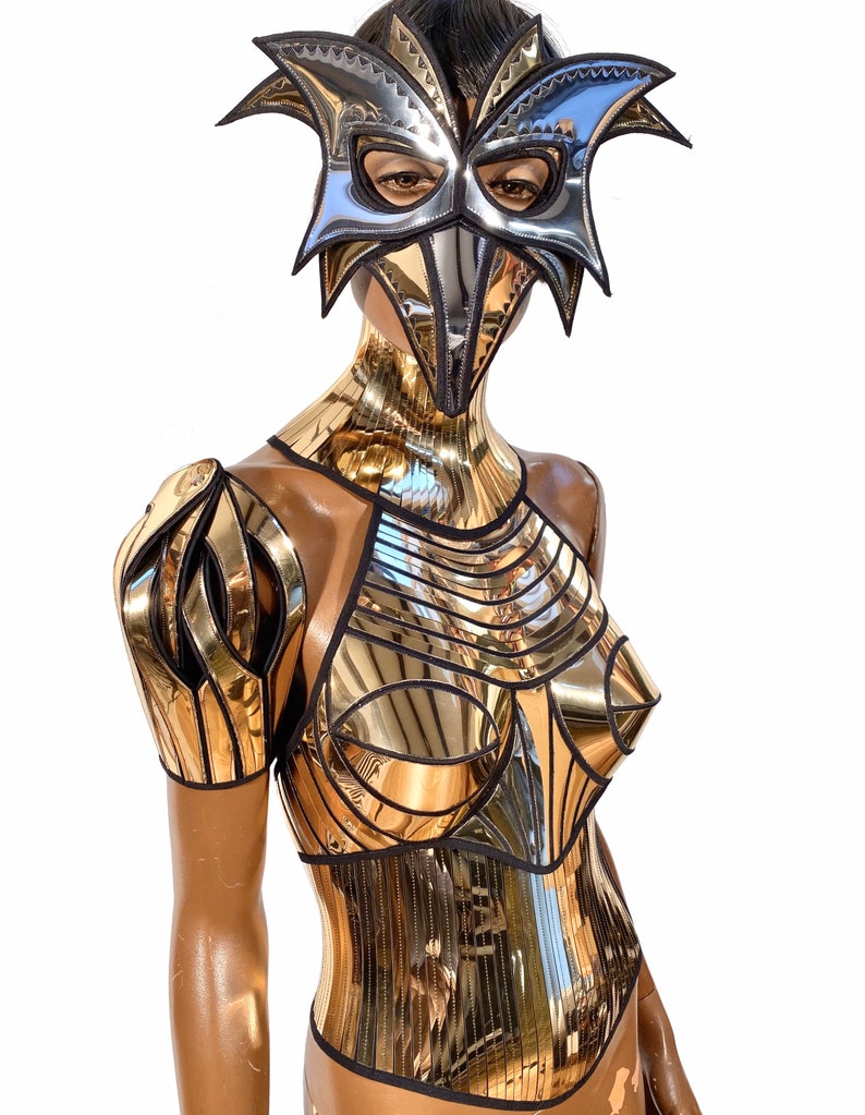 May include: A gold and silver mirrored costume with a mask. The costume features a halter top, a corset, and shoulder pads. The mask is a bird-like design with a long beak.