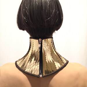 Op de afbeelding: Een gouden en zwarte choker met een ritsluiting. De choker is gemaakt van een stof met een golvende textuur.