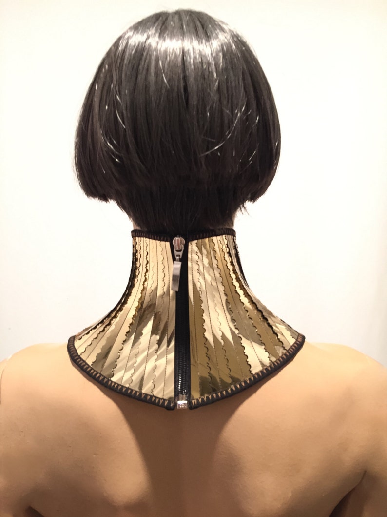 Op de afbeelding: Een gouden en zwarte choker met een ritsluiting. De choker is gemaakt van een stof met een golvende textuur.