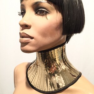 Op de afbeelding: Een gouden, gespiegelde choker ketting met een zwarte rand. De choker is gemaakt van meerdere verticale stroken gespiegeld materiaal, waardoor een geometrisch patroon ontstaat.