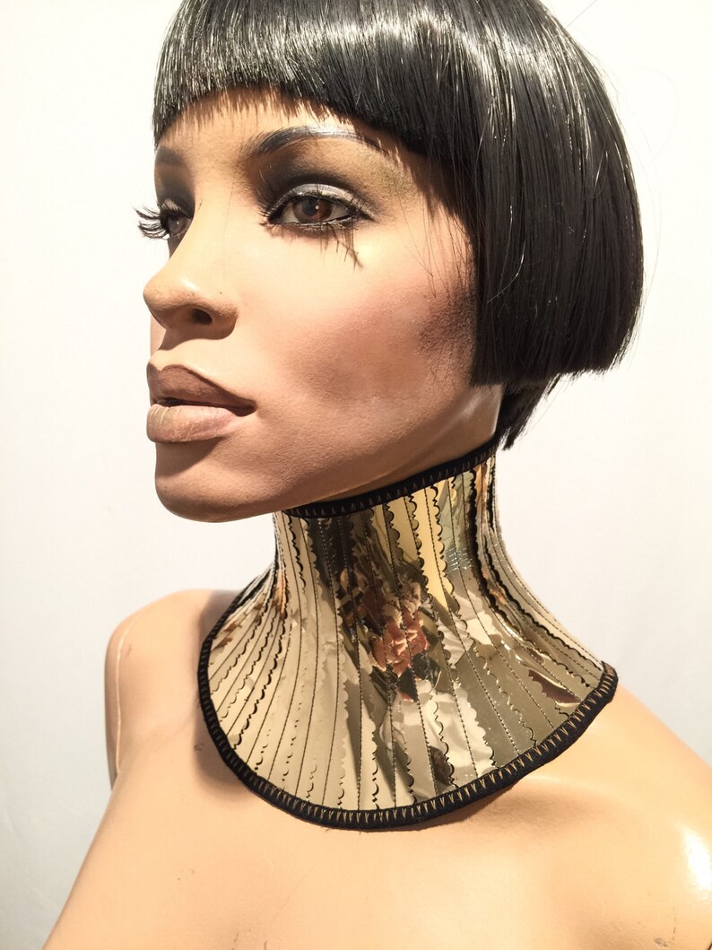 Op de afbeelding: Een gouden, gespiegelde choker ketting met een zwarte rand. De choker is gemaakt van meerdere verticale stroken gespiegeld materiaal, waardoor een geometrisch patroon ontstaat.