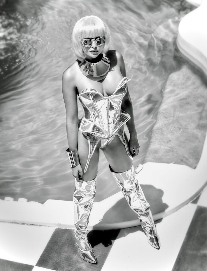 May include: A woman wearing a silver, metallic corset with a silver choker necklace and silver thigh-high boots. She is standing in front of a pool.