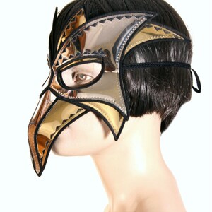 May include: A gold and silver masquerade mask with a long, pointed beak. The mask is made of leather and has a black elastic strap.