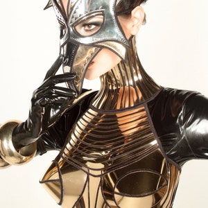 May include: A woman wearing a black latex bodysuit with a gold metallic bodice and a silver and gold mask. The mask is shaped like a bird's head and covers the woman's eyes and nose.