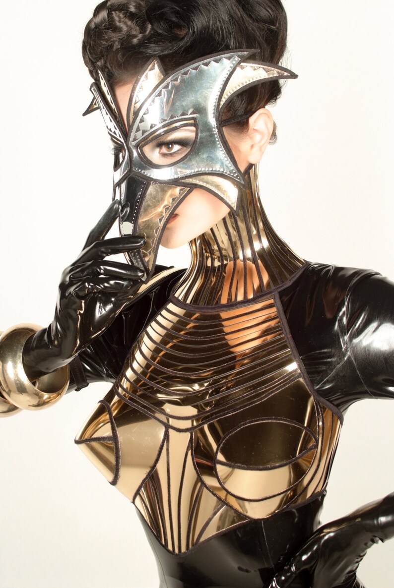 May include: A woman wearing a black latex bodysuit with a gold metallic bodice and a silver and gold mask. The mask is shaped like a bird's head and covers the woman's eyes and nose.