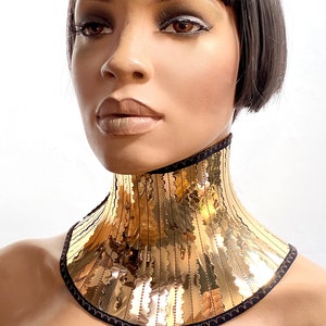 Op de afbeelding: Een gouden metalen choker ketting met een zwarte fluwelen achterkant. De ketting heeft een golvende, gekrulde rand en is gemaakt van veel dunne, verticale metalen stroken.