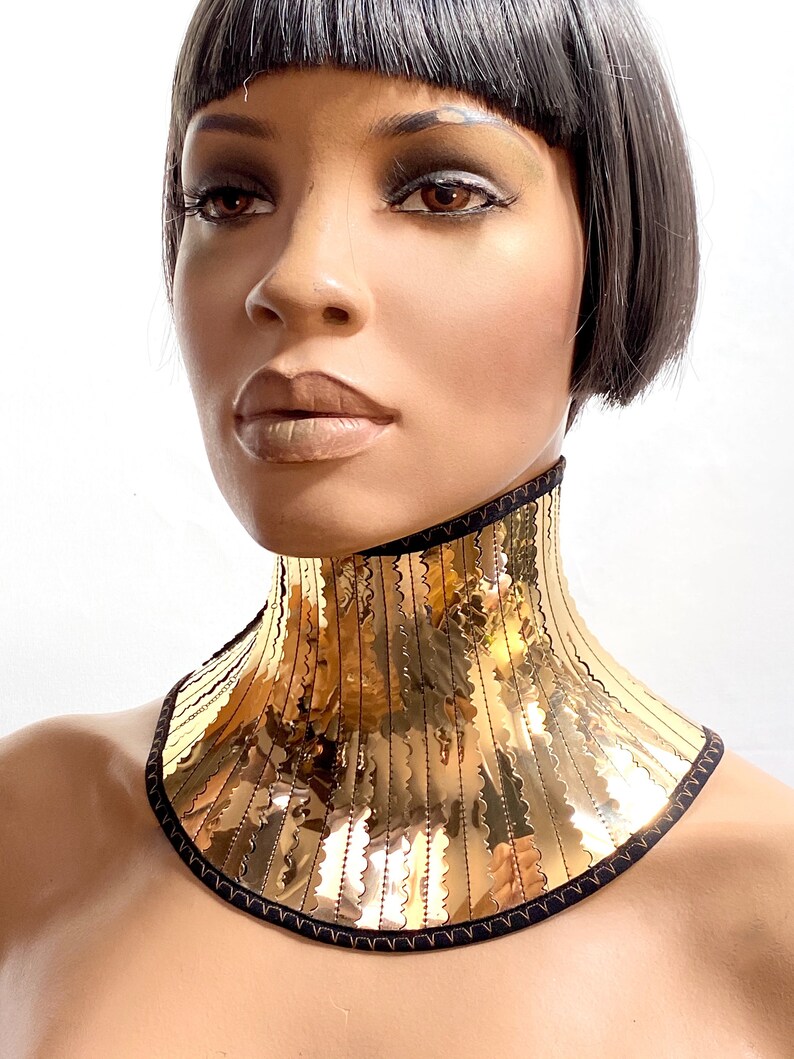 Op de afbeelding: Een gouden metalen choker ketting met een zwarte fluwelen achterkant. De ketting heeft een golvende, gekrulde rand en is gemaakt van veel dunne, verticale metalen stroken.