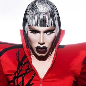 May include: A person wearing a red jacket with black accents and a silver wig. The person has dark makeup and a serious expression.