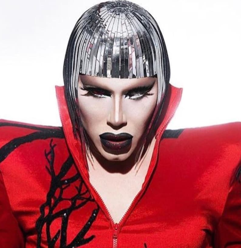 May include: A person wearing a red jacket with black accents and a silver wig. The person has dark makeup and a serious expression.