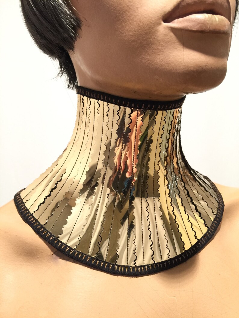 Op de afbeelding: Een gouden chokerketting met een gegolfde rand en zwarte bies. De ketting is gemaakt van metaal en heeft een reflecterend oppervlak.