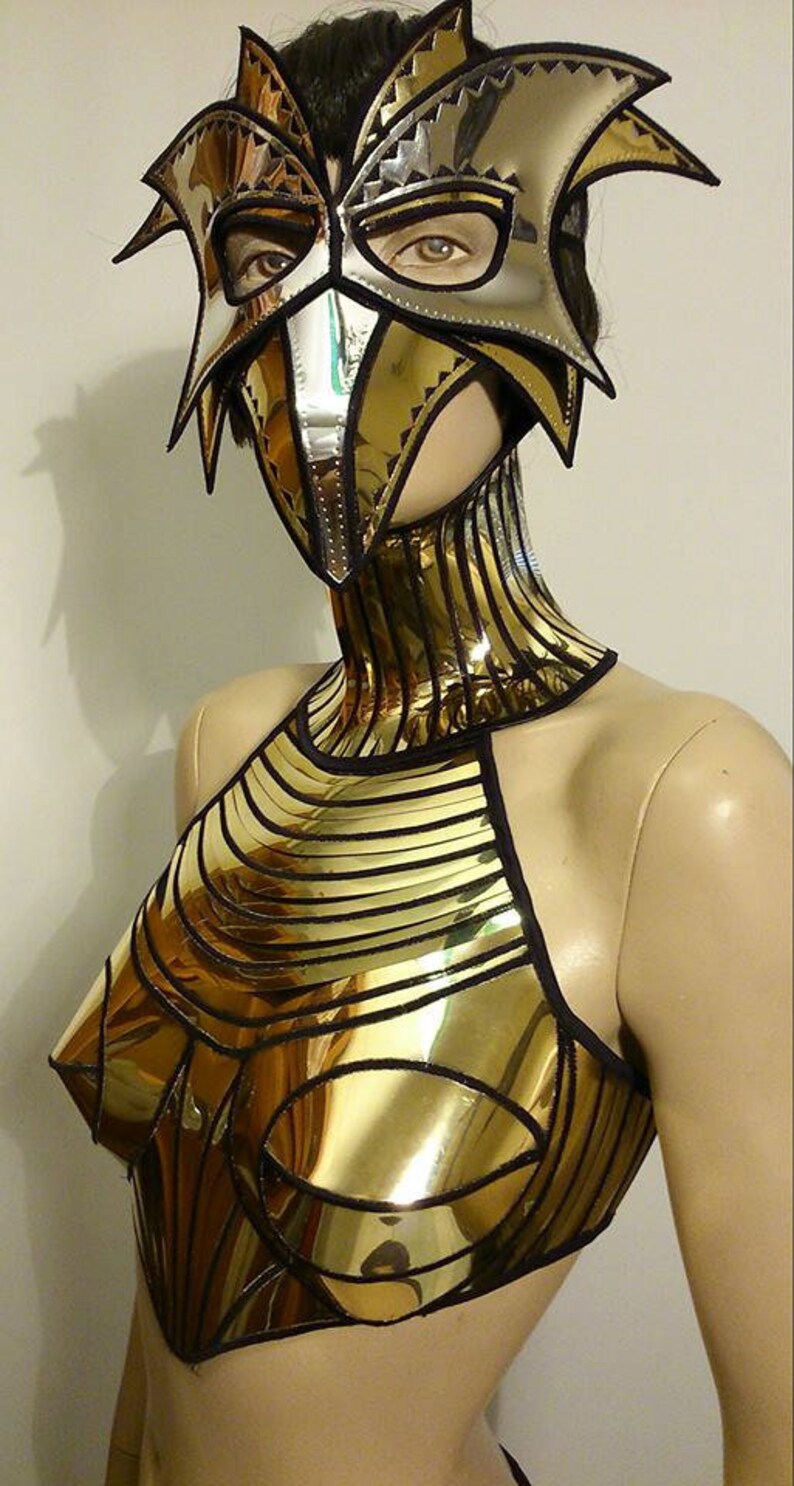 May include: A gold and silver metallic mask with a bird-like design. The mask is worn by a mannequin with a gold and black metallic bodice.