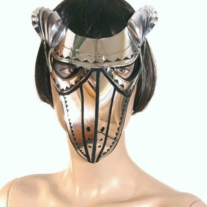 May include: A silver and black masquerade mask with a pointed beak and decorative details. The mask has a mirrored surface and is worn by a person with black hair.