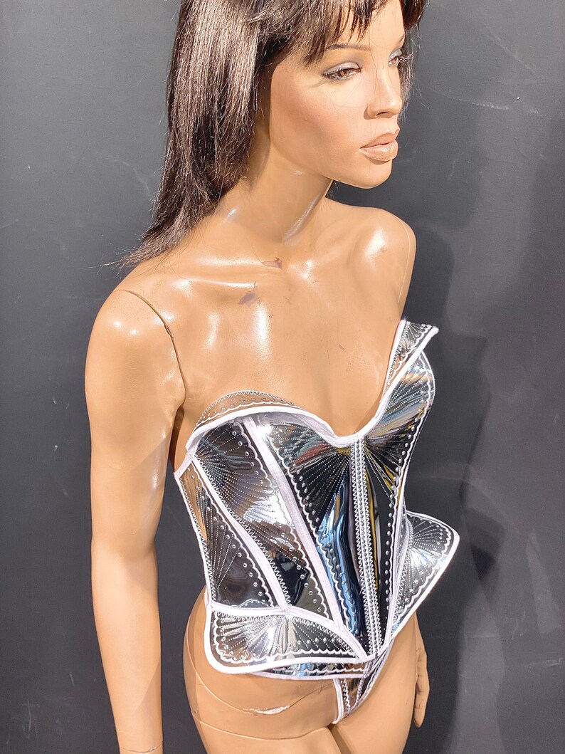 May include: A silver and white corset with a decorative design. The corset is made of a shiny, reflective material and has a white trim.