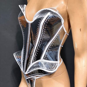 May include: A silver and white corset with a structured design and a scalloped edge. The corset is made of a shiny, reflective material and has a white trim.