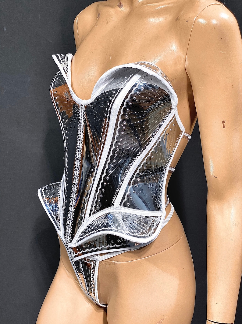 May include: A silver and white corset with a structured design and a scalloped edge. The corset is made of a shiny, reflective material and has a white trim.