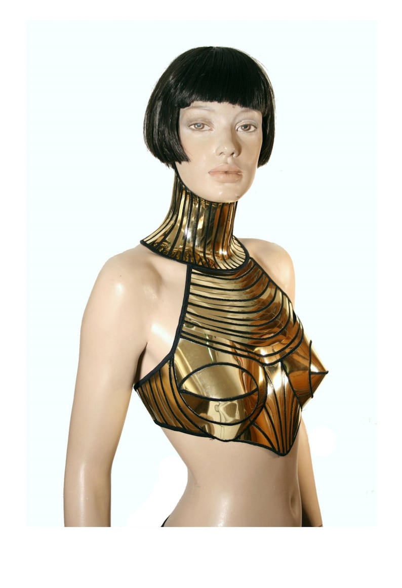 May include: A metallic gold chest piece with a high neck collar and structured conical bust cups. The garment features black piping along the edges and between the segmented panels, creating a rigid, reflective surface that covers the upper torso.