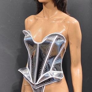 May include: A silver and white corset with a white trim and a heart-shaped neckline. The corset has a shiny, metallic finish and a fitted silhouette.