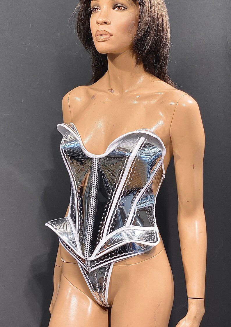 May include: A silver and white corset with a white trim and a heart-shaped neckline. The corset has a shiny, metallic finish and a fitted silhouette.