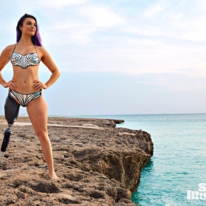 May include: A woman wearing a silver and black bikini stands on a rocky cliff overlooking the ocean. She has purple hair and is wearing a prosthetic leg.