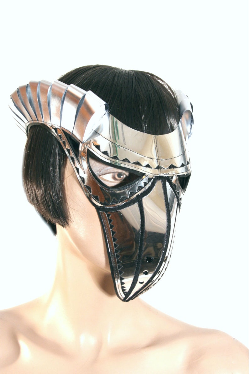 May include: A silver and black masquerade mask with a pointed beak and a decorative design. The mask is worn by a mannequin with black hair.