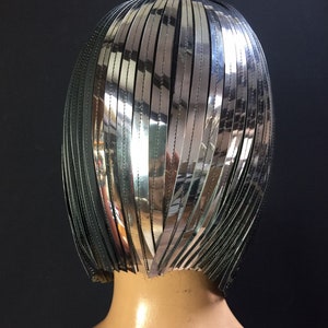 May include: A silver and black wig with a short, straight style. The wig is made of many thin strips of silver material that are sewn together.