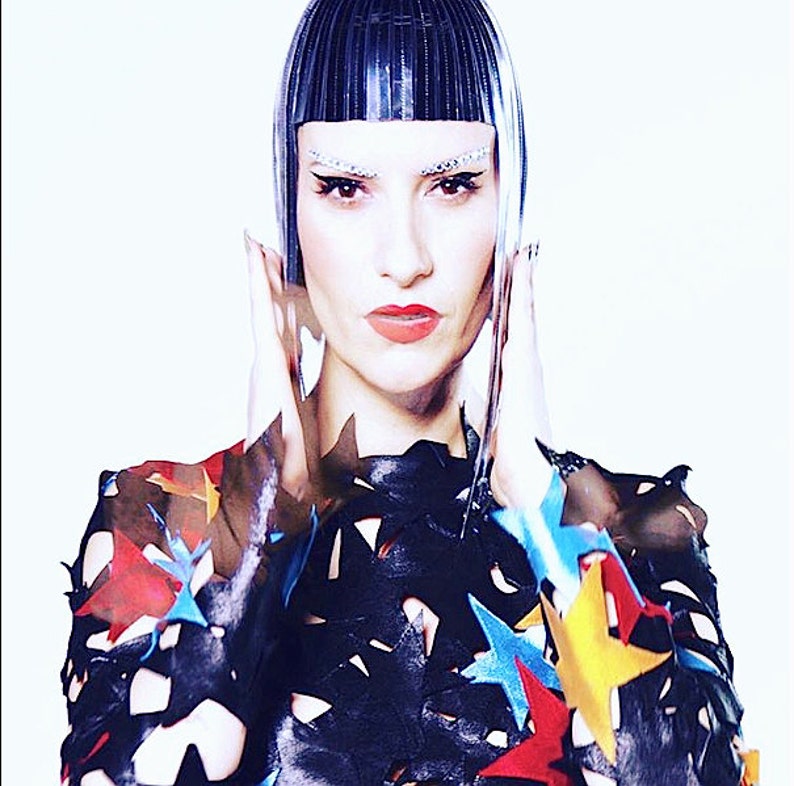 May include: A woman with a black and silver geometric hairstyle wears a black leather top with colorful star cutouts.