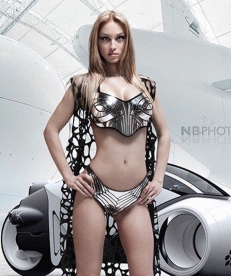 May include: A woman wearing a silver and black metallic bikini top and matching bottoms. She is standing in front of a white futuristic car. The car has a large wheel on the right side. The image is in a studio setting with a white background.