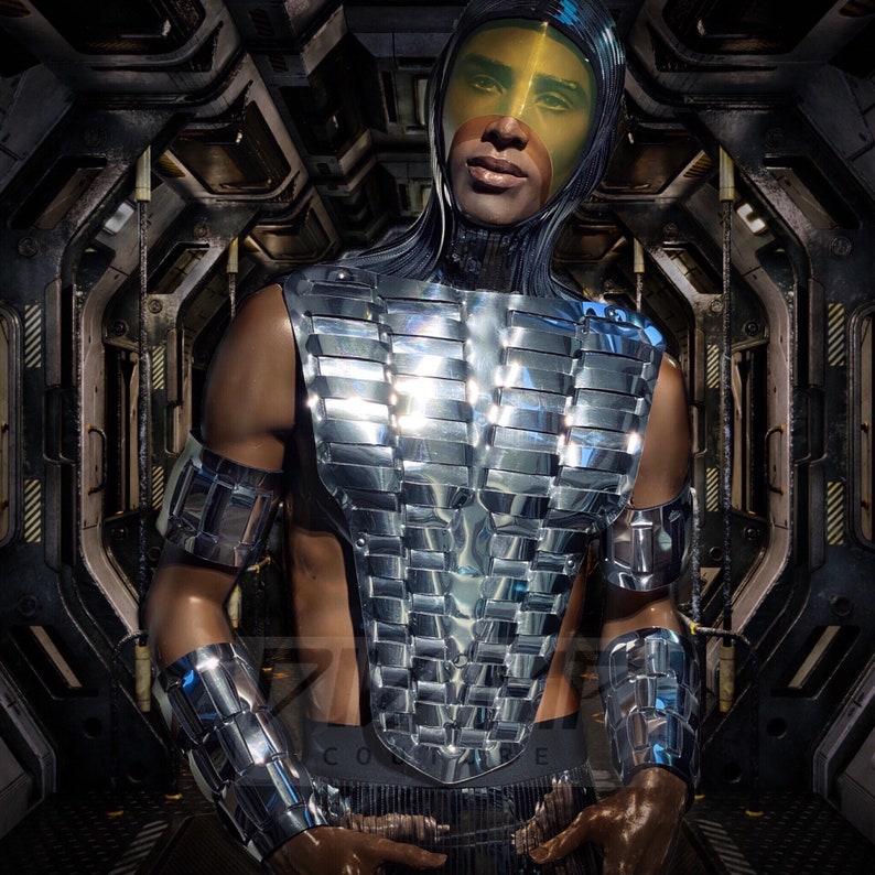 Pu&ograve; includere: Una persona che indossa un costume futuristico argentato, segmentato, con un casco giallo. Il costume ha un design geometrico ed &egrave; riflettente.