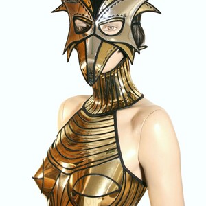 May include: A gold and silver metallic mask with a bird-like design. The mask is worn by a mannequin with a gold metallic top and a matching gold metallic collar.