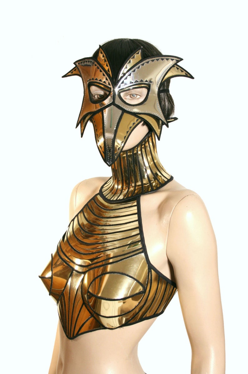 May include: A gold and silver metallic mask with a bird-like design. The mask is worn by a mannequin with a gold metallic top and a matching gold metallic collar.