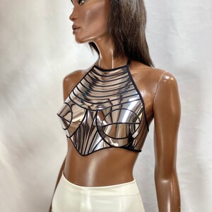May include: A metallic silver halter top featuring a reflective, mirror-like finish with structured, moulded cups and black trim detailing. The garment has a high neckline and a cropped, pointed hemline, constructed from segmented panels.