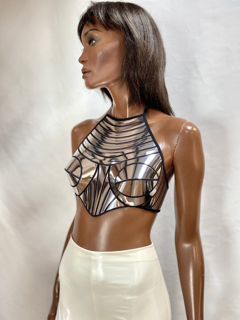 May include: A metallic silver halter top featuring a reflective, mirror-like finish with structured, moulded cups and black trim detailing. The garment has a high neckline and a cropped, pointed hemline, constructed from segmented panels.