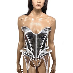 May include: A silver and white corset with a white lace trim. The corset has a sweetheart neckline and a boned structure. The corset is paired with a matching garter belt and stockings.