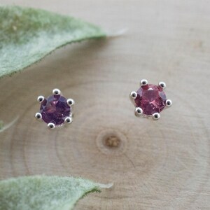Peut inclure: Une paire de boucles d'oreilles à clous avec des pierres précieuses rondes et facettées d'un rose profond, serties dans des griffes en métal argenté. Chaque boucle possède un sertissage à six griffes qui maintient la pierre circulaire.
