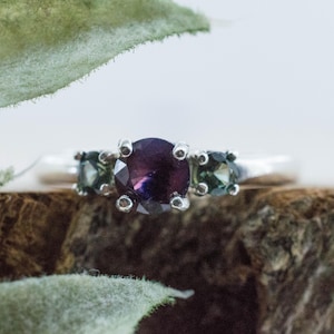 Könnte beinhalten: Ein Silberring mit einem großen, runden, tiefvioletten Mittelstein, der von zwei kleineren, runden, dunkelgrünen Seitensteinen flankiert wird. Die Steine werden von Silberfassungen auf einer glatten, polierten Silberschiene gehalten.